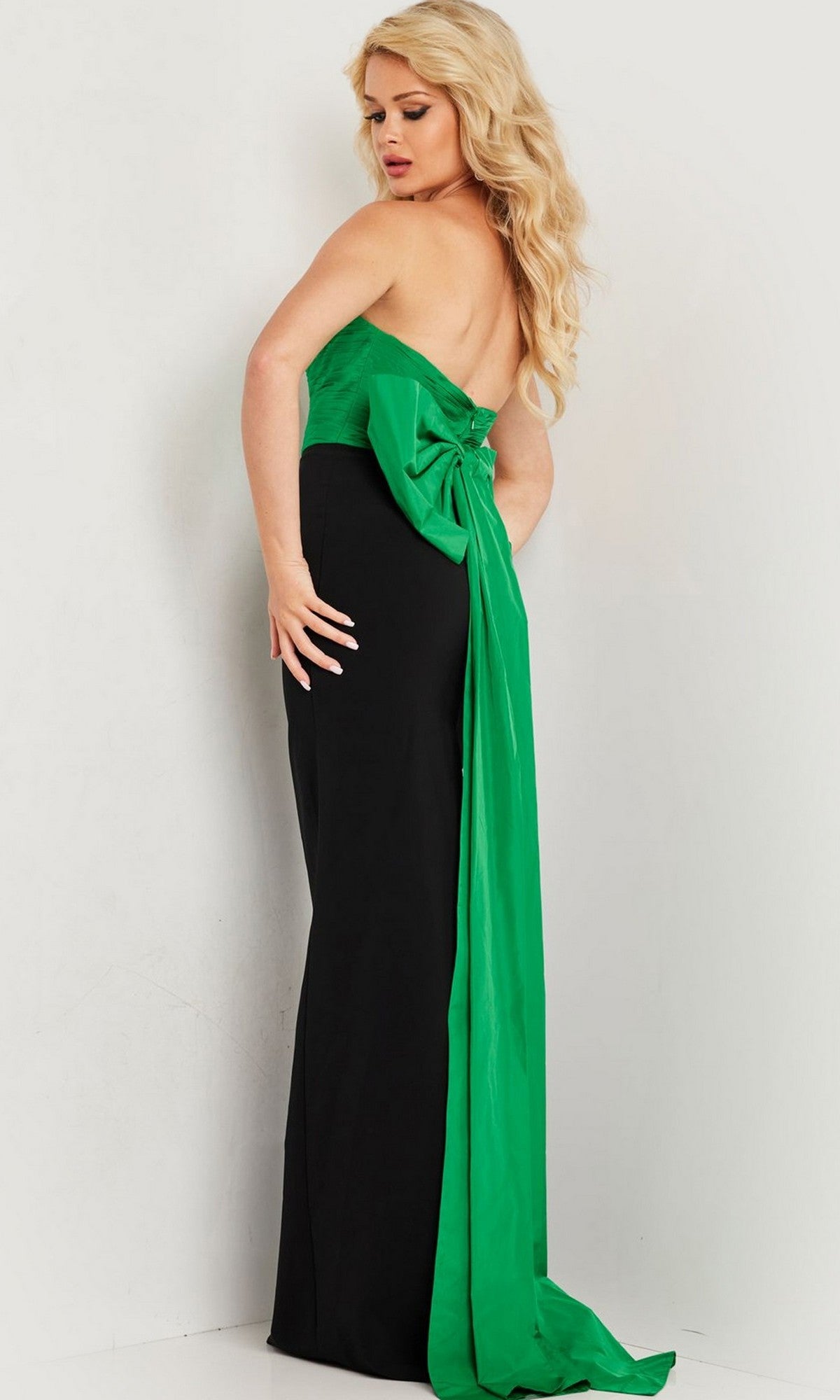 A woman stands against a light background, wearing the Jovani 36670 Long Prom Dress—strapless, black, and featuring a striking green bow and sash detail on the back.